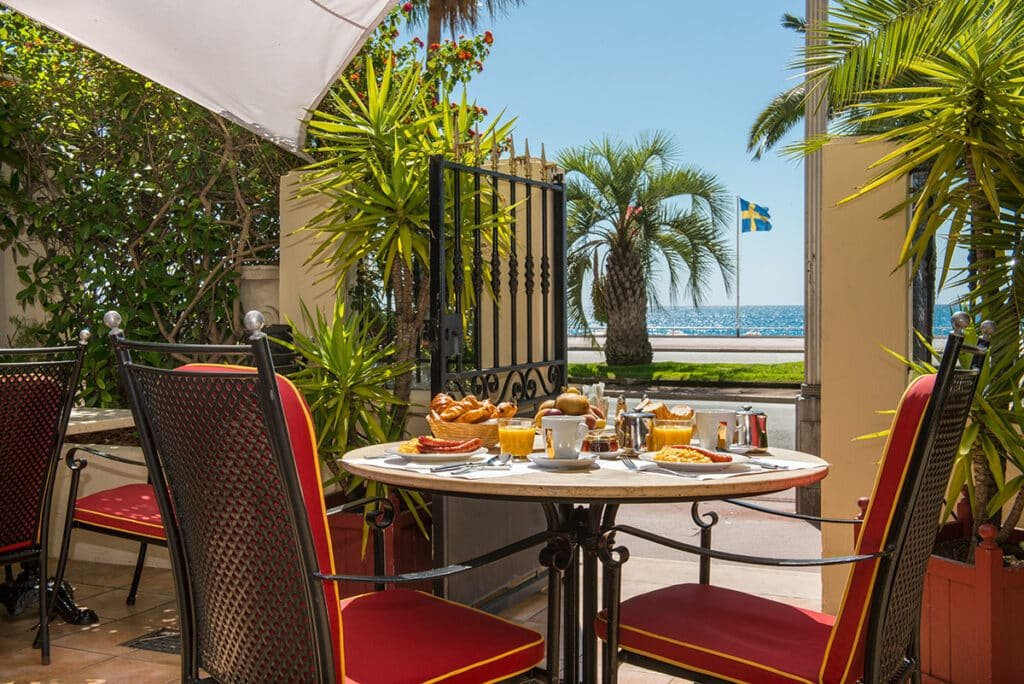 Área de refeições externa do Hôtel West End Promenade. Há uma mesa redonda com duas cadeiras em volta. Em cima da mesa há itens de café da manhã como xícaras de café, copos de suco, cesta de pães e pratos com ovos. O local é cercado por um muro com plantas e tem vista e uma porta que dá acesso a avenida beira-mar.