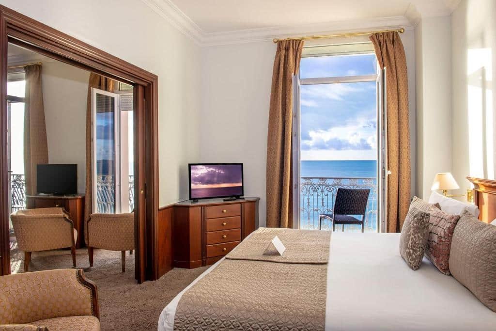 Quarto do Hôtel West End Promenade. O quarto é amplo e tem uma decoração clássica. A cama está encostada na parede do lado direito, ao lado há uma porta para a varanda com uma cadeira e vista para o mar e ao lado uma cômoda com TV. No lado esquerdo do quarto há uma passagem para outra área com duas poltronas, uma outra cômoda com TV e duas portas para a varanda. Imagem para ilustrar o post hotéis em Nice.