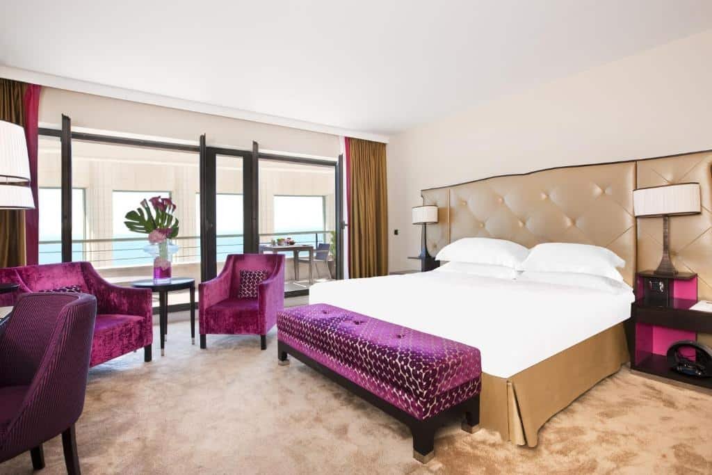 Quarto do Hyatt Regency Nice Palais de la Méditerranée. A cama está encostada na parede do lado direito do quarto, entre a cama há duas cômodas pequenas com abajour e em frente a cama a uma mesa, cadeira, mesa de centro e duas poltronas. A parede esquerda do quarto é de vidro e dá acesso a uma enorme varanda com mesa e cadeiras. Imagem para ilustrar o post hotéis em Nice.