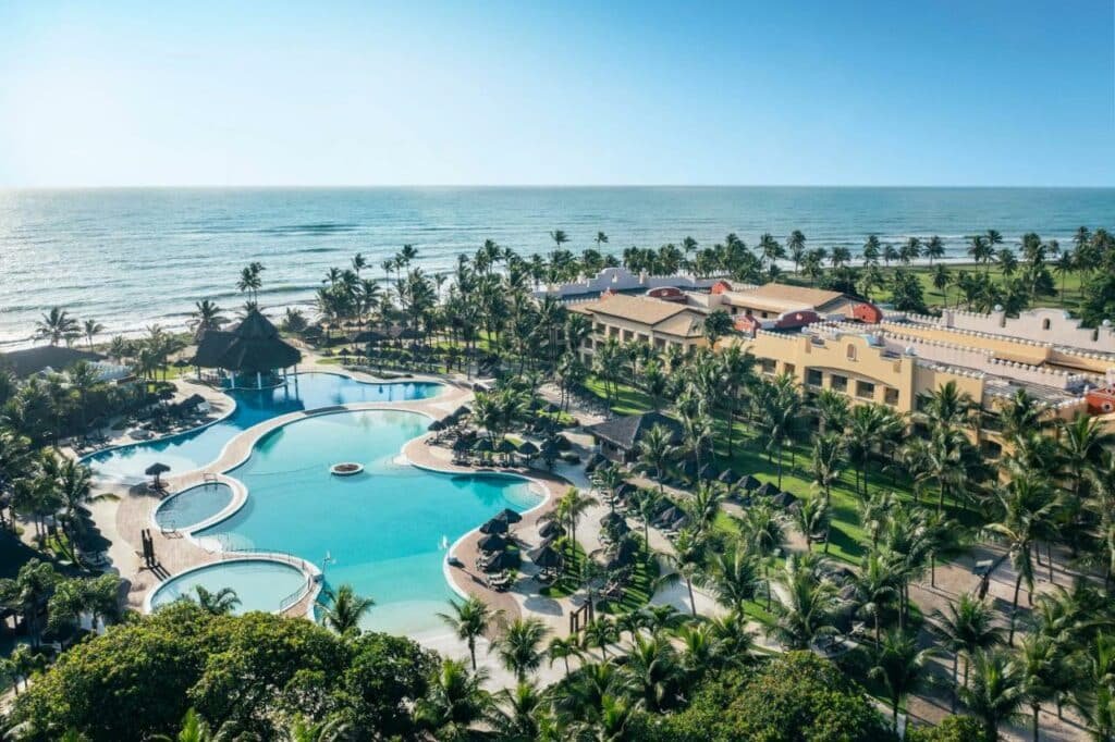 Vista de cima do resort Iberostar Bahia - All Inclusive. É possíver ver piscinas de diferentes tamanhos e formatos do lado esquerdo da imagem. Já do lado direito está o próprio resort. Existem muitas árvores em toda a imagem e a praia ao fundo.