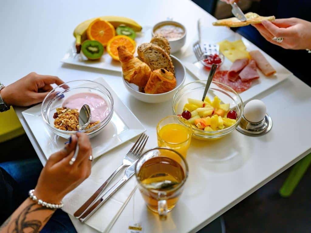 Foto de um café da manhã sendo realizado por duasa pessoas no ibis budget Hotel Luzern City. A mesa é branca e vemos apena as mãos de duas mulheres, que estão sentadas de frente uma para a outra. A da esquerda come uma tigela com iogurte e cereais, e ao seu lado há um copo com chá e suco. Já a da direita está comendo um prato com frios, e ela passa geleia em uma torrada. No centro da mesa há mais opções em tigelas que contém frutas, pães e massas.