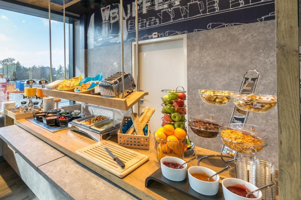 Buffet no restaurante do ibis budget Geneve Palexpo Aeroport. O ambiente está iluminado pois possui uma grande janela à esquerda. A bancada de pedra contém diversas opções de frutas, geleias, cereais, pães, etc,