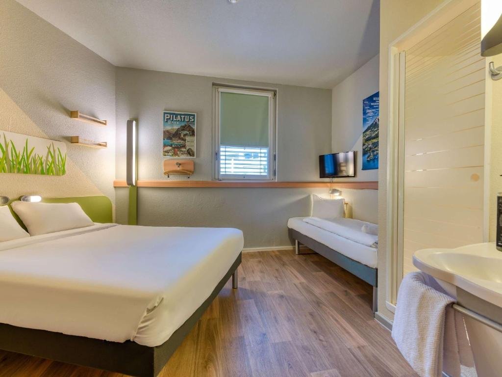 Foto do ibis budget Hotel Luzern City, para representar o post de hotéis em Lucerna. O quarto possui um espaço agradável. A cama ade casal está na esquerda, encostada na parede. Na sua frente há um closet. Do lado do closet, no canto inferior direito da foto, existe uma pia, que vemos parcialmente. Há um mini sofá no fundo do quarto, uma TV voltada para a cama e uma janela pequena.
