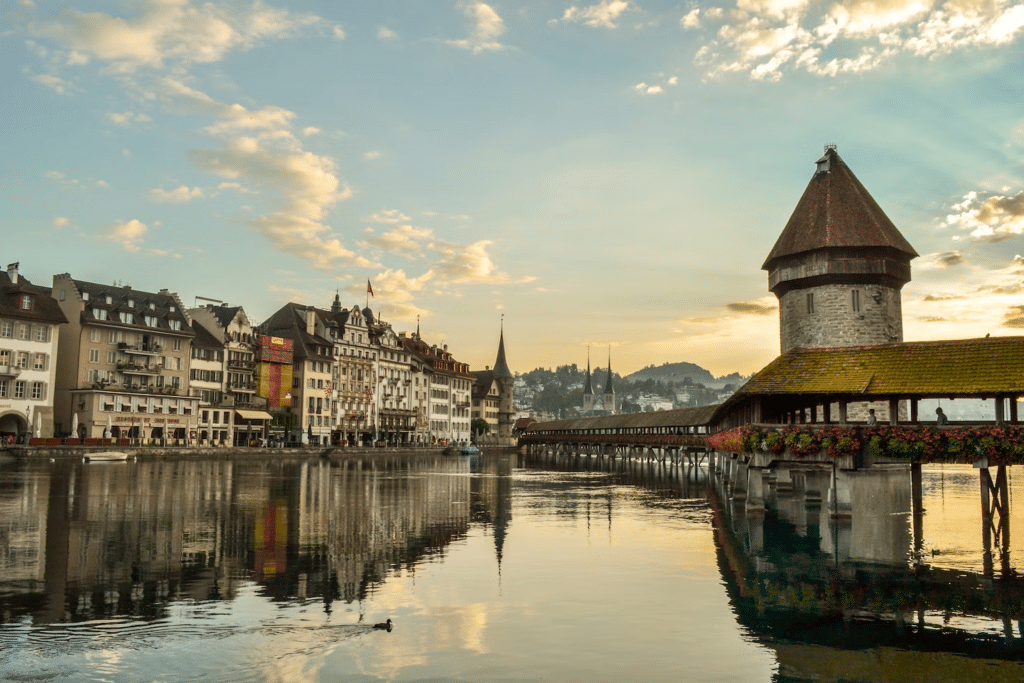Foto tirada de dia da cidade de Lucerna. A Ponte da Capela está à direita, e ao fundo vemos as mais diversas construções históricas no centro. Abaixo da ponte vemos o Rio Reuss, que reflete as nuvens e os prédios.