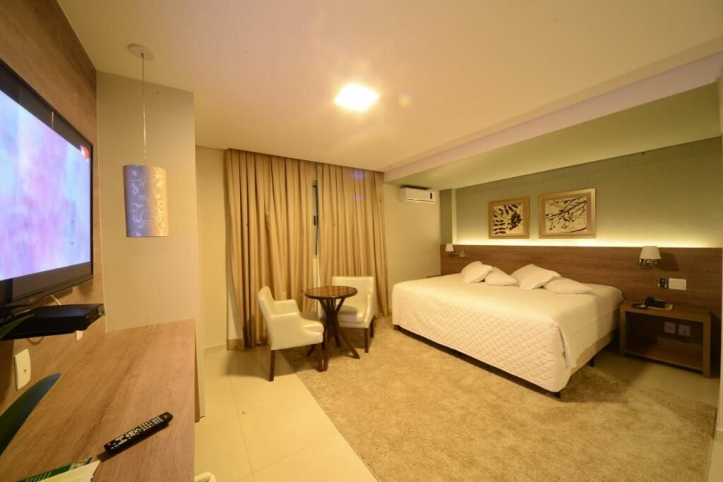 Quarto com uma cama grande de casal. luz amarela, janela com corinas e uma mesa redonda com duas cadeiras. No canto da imagem há uma televisão.