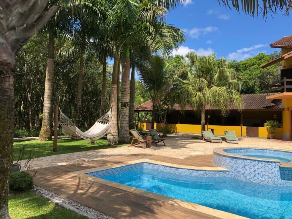 Área da piscina da acomoação Jardim Boêmio. A piscina está do lado direito da foto. Do lado esquerdo estão algumas cadeiras, espreguiçadeiras e uma rede pendurada entre duas árvores. Ao fundo há uma área coberta da pousada.