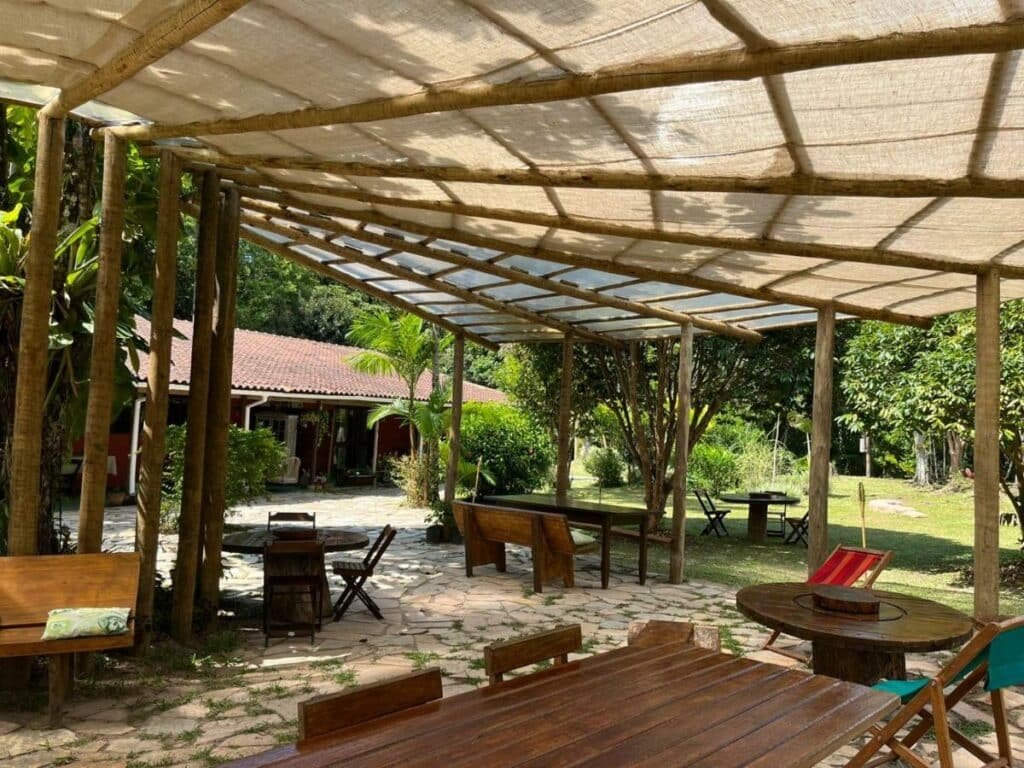 Área comum da pousada Jardim Mirim. Ela é coberta e está rodeada por árvores, plantas e grama. Existem várias mesas, cadeiras e bancos. Ao fundo é possíver ver uma parte da pousada.