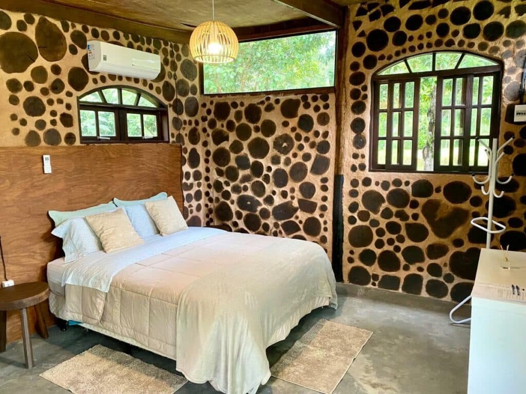 Quarto da pousada Jardim Mirim. Uma cama de casal está no canto perto da parede. Um banquinho está ao seu lado e, em frente, uma mesa. Existem duas janelas na parede ao lado da cama e uma pequena janela acima dela.