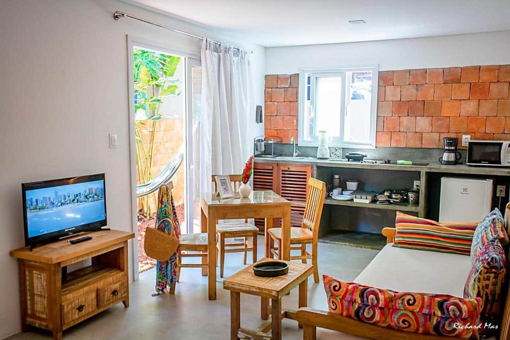 Sala de estar e cozinha do Jardim Secreto. Do lado direito um sofá cama, no meio uma mesa de centro, do lado esquerdo uma raque com televisão e uma mesa de madeira. No fundo uma mini cozinha.