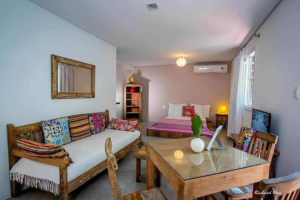 Flat do Jardim Secreto. Do lado direito na frente uma mesa de madeira, do lado esquerdo um sofá cama com um espelho em cima. No fundo uma cama de casal, em cima um ar-condicionado.