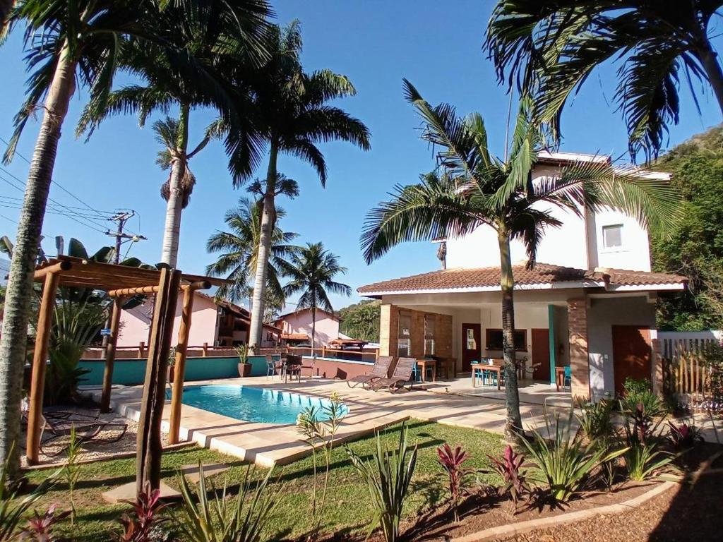 Propriedade do Kauano Pousada Barequeçaba com uma piscina em um jardim com gramado e palmeiras, o pequeno deck ainda tem espreguiçadeiras e uma parte coberta com uma mesa e cadeiras