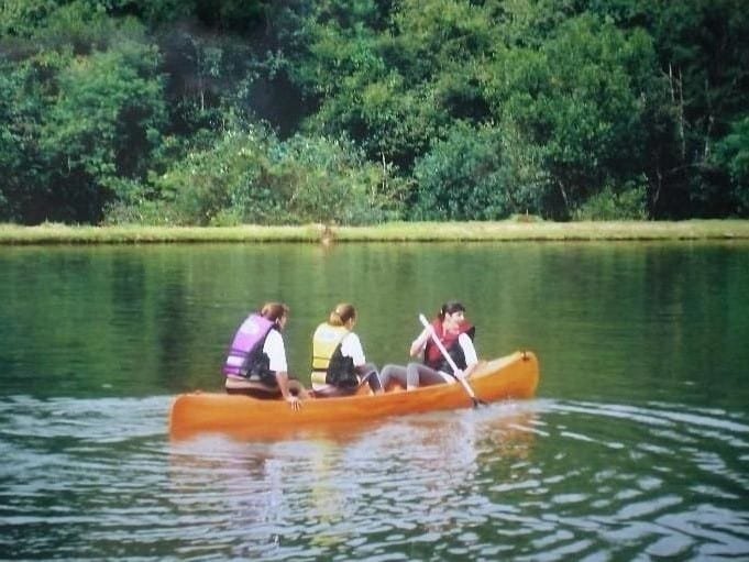 A imagem mostra três pessoas em um barco de canoagem laranja, remando em um lago cercado por vegetação densa e árvores verdes no Leão de Judá Charme Hotel. Todos usam coletes salva-vidas, e cada um segura um remo enquanto avançam calmamente pela água tranquila. O ambiente parece ser natural e sereno, com o barco criando leves ondulações na superfície do lago. Ao fundo, a margem do lago é verde e coberta de vegetação, reforçando o clima de tranquilidade da cena.