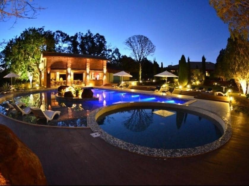 A imagem mostra uma bela área de lazer ao ar livre em um ambiente luxuoso, com uma piscina iluminada à noite. A piscina principal tem formato irregular e está cercada por espreguiçadeiras brancas dispostas em um deck de madeira. Há uma piscina menor e redonda conectada à maior. Ao fundo, há uma construção de tijolos aparentes com luzes aconchegantes, além de um bar coberto e algumas mesas. Árvores e arbustos cuidadosamente podados complementam o cenário tranquilo e acolhedor. Esse é o Leão de Judá Charme Hotel, um dos hotéis-fazenda em Minas Gerais.