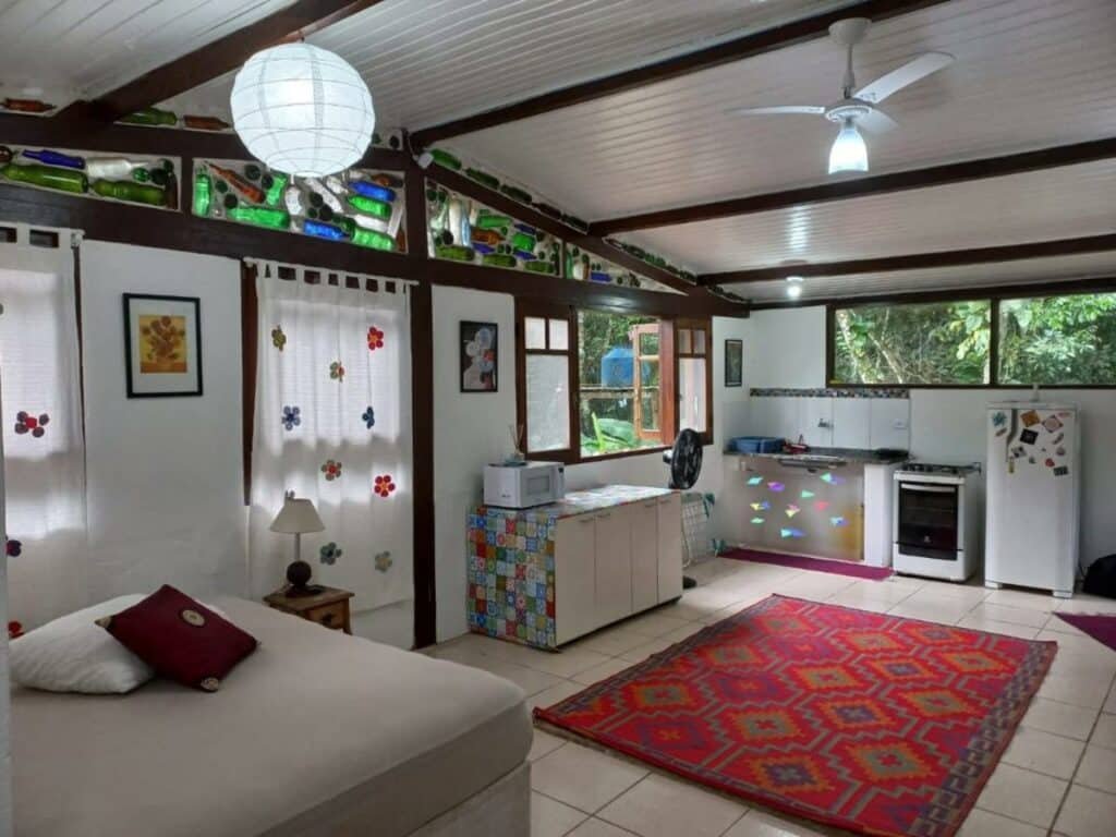 Interior do Loft dos Vagalumes. Do lado direito está a cozinha com uma pia, um fogão e uma geladeira encostados na parede. Em frente a eles está um armário e um grande tapete no chão. Do lado esquerdo está uma cama de casal.