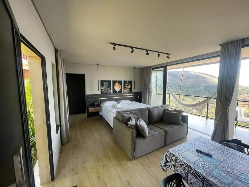 área interna do Loft Paraíso da Serra com uma cama ao centro, três quadros de fundo preto com plantas desenhadas pendurados em cima da cabeceira, um sofá cinza disposto em frente aos pés da cama, e ao lado direito é possível enxergar a varanda ampla com vista para o vale e com uma rede branca pendurada