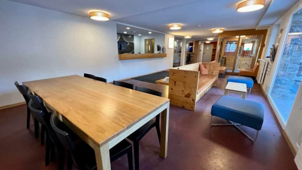 Lounge compartilhado do Zermatt Youth Hostel com uma mesa quadrada do lado esquerdo da imagem com oito cadeiras e ao fundo bancoos,