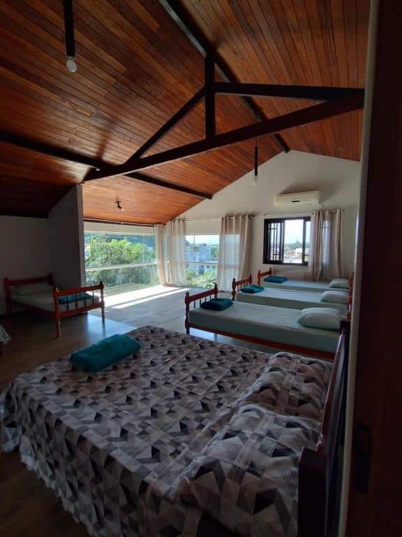 Quarto do Mandallah Hostel com uma cama de casal e outras quatro camas de solteiro dispostas lado a lado. O quarto tem teto alto de madeira e uma área, ao fundo, com amplas janelas e muita luz natural, onde é possível enxergar a cidade