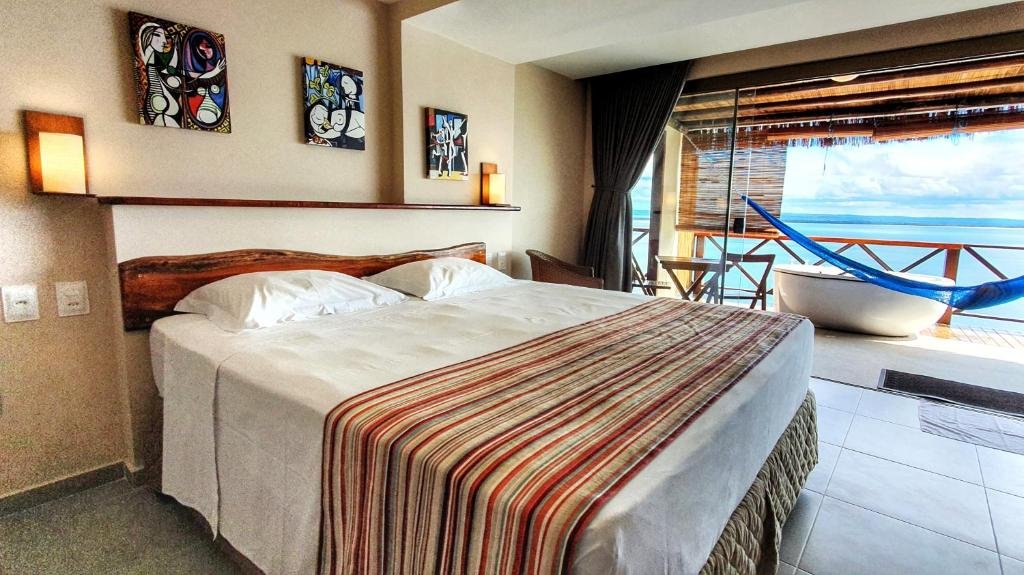 Quarto do Mar e Morro Flats. Uma cama de casal do lado esquerdo, no fundo, a varanda com mesa e cadeiras, uma rede e uma banheira de hidromassagem com vista para o mar.