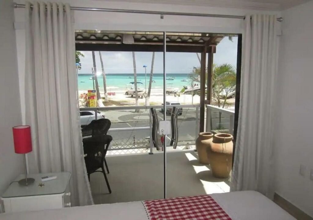 Parte da cama do Maragogi Beach Flats, com uma porta de vidro dando acesso à uma sacada com cadeiras pretas, dois vasos e vista da praia que é dividida apenas por uma rua curta.