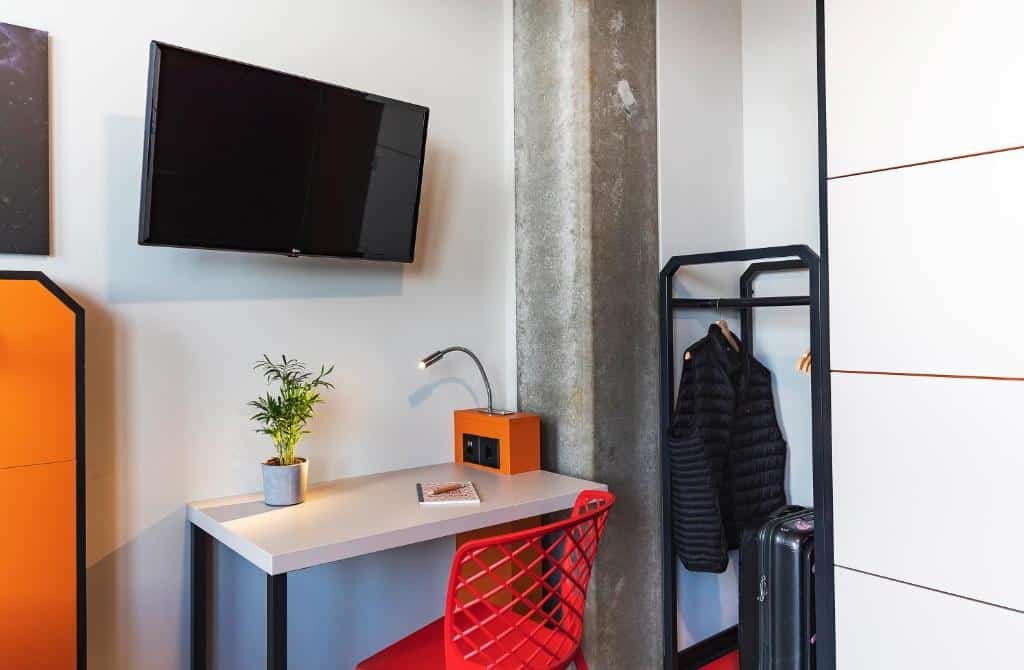 Outro ângulo na suíte do Meninger Hotel Geneve. Vemos uma escrivaninha com abajur e um vaso de plantas em cima, acompanhada de uma cadeira vermelha. Em cima, há uma TV fixa na parede, e à direita, a arara de roupas.