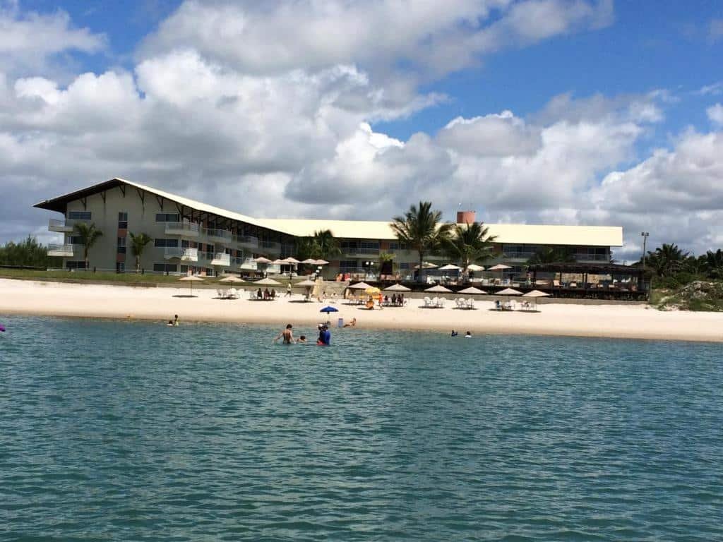 Praia com algumas pessoas nadando, uma faixa de areia com alguns guarda-sóis e ao fundo o Muro Alto Suites – Marupiara, um dos hotéis em Muro Alto