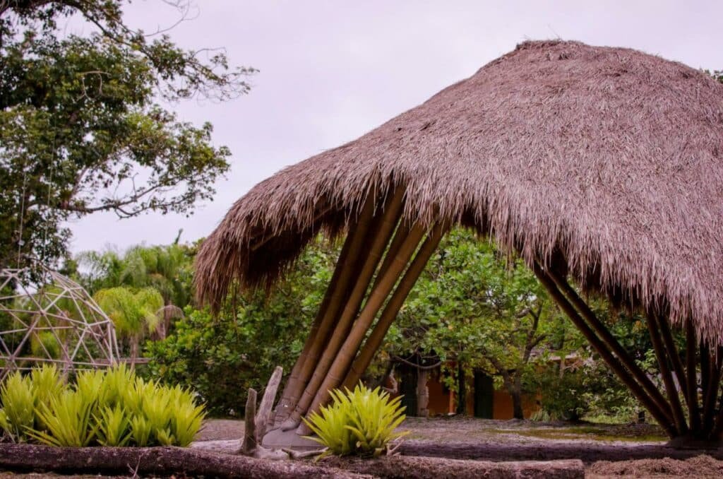 Área de fora da Namastê Pousada e Camping. Em primeiro plano há uma tenda de palha, perto de açgumas árvores  plantas. Em segundo plano é possível ver a própria pousada.