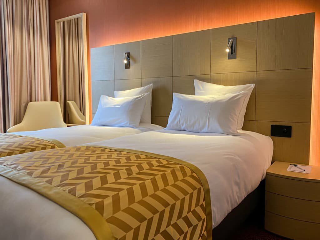 Foto de um quarto do Nash Airport Hotel, para ilustrar o post de hotéis em Genebra. Vemos duas camas juntas, com lençóis brancos e uma cabeceira de madeira atrás, que contém duas luminárias e tomada. Do lado esquerdo, vemos uma poltrona e parte da cortina.