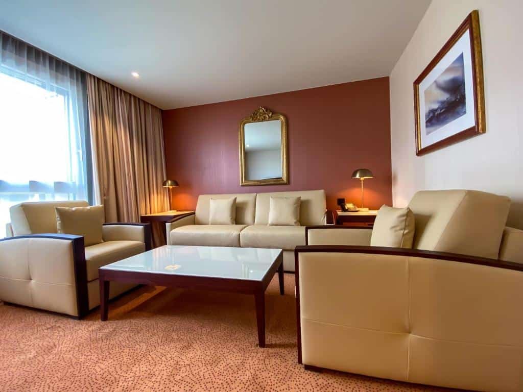 Uma sala no Nash Airport Hotel. Vemos três sofás. Um está encostado na parede no fundo, no centro, e os outros dois estão de lado. No meio dos três, há uma mesa quadrada. A janela do quarto está na parede esquerda, atrás de um dos sofás.