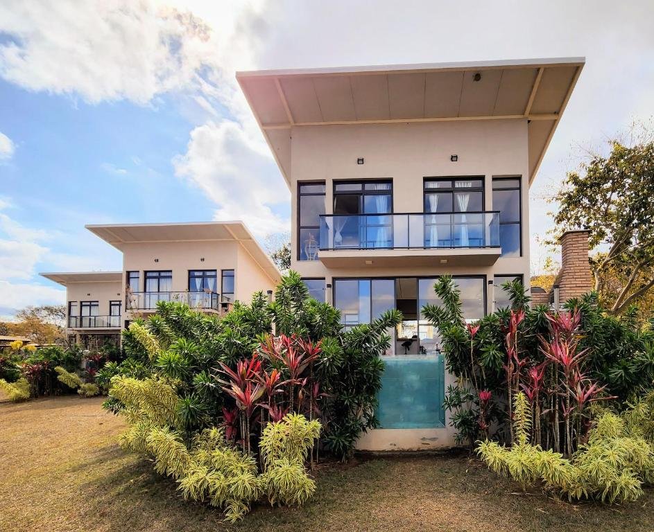 fachada frontal das casas Nativos de Minas. São três casas retangulares enfileiradas lado a lado, iguais umas as outras, com varanda aberta e muitas portas e janelas de vidro. Em frente a casa há belas flores e plantas no jardim