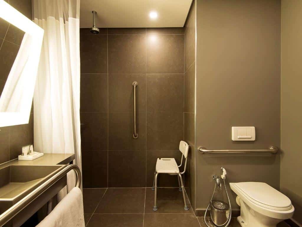 Banheiro adaptado do Novotel Santos Gonzaga com barras de apoio, box sem vidro, uma cadeira perto do chuveiro e outras adaptações