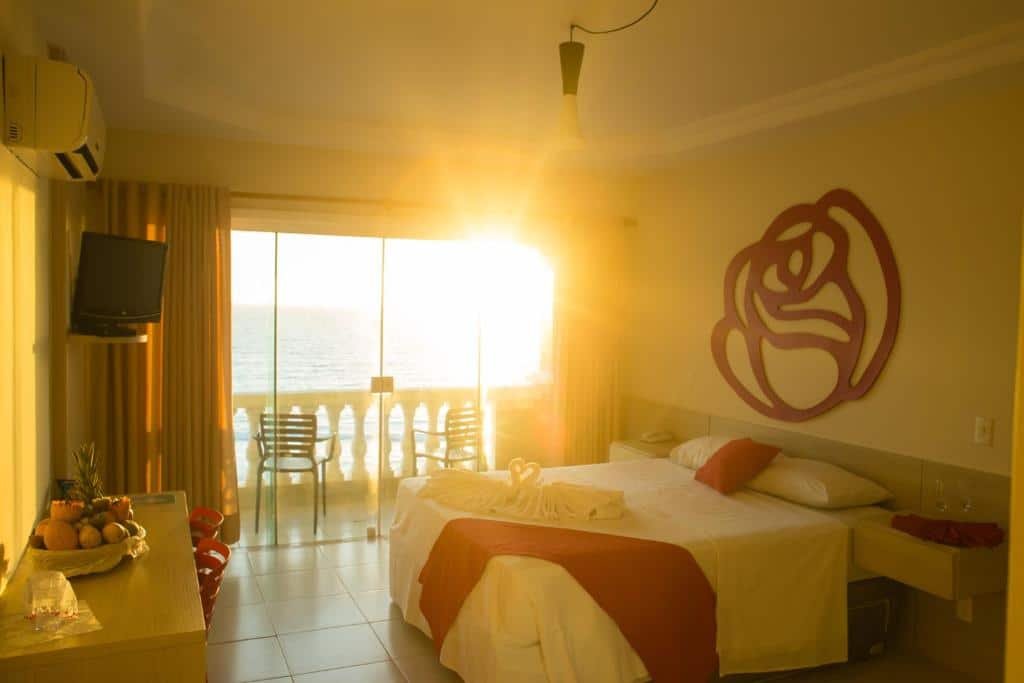 Quarto do Ocean View Hotel. No lado esquerdo do quarto há uma mesa com duas cadeiras e uma TV presa na parede. Já no lado direito está a cama de casal e no fundo há uma porta de vidro que dá acesso a varanda. A varanda tem vista total para o mar e também possui duas cadeiras.