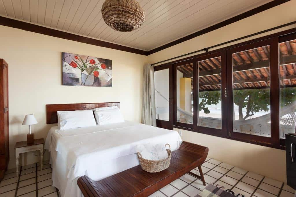 Quarto do hotel oidá maragogi, com uma cama de casal, uma mesinha ao lado com um abajur, na frente da cama tem um banco com um cesto com toalhas enroladas dentro e, à direita, tem uma janela mostrando a varanda com rede, uma árvore e o mar ao fundo.