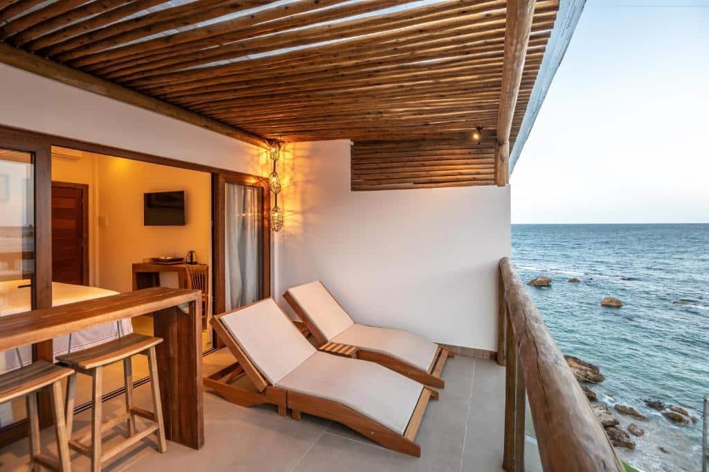 Varanda do One Beach Boutique Hotel. Do lado direito o mar, No meio cadeiras de tomar sol, uma banqueta com cadeiras. Do lado esquerdo o quarto.