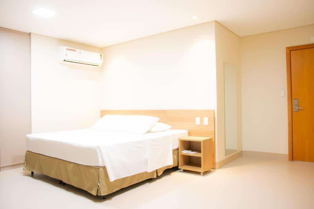 Quarto com uma cama de casal, mesinha ao lado, espelho e porta no canto. É possível ver também um ar-condicionado.