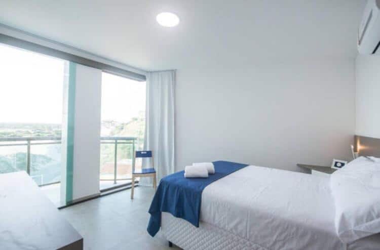 Quarto do airbnb Orla Praia Grande. No canto direito da imagem há uma cama de casal e uma pequena mesa no lado esquerdo da cama. Já no lado esquerdo da imagem é possível ver parte de uma outra mesa e no fundo há uma grande porta de vidro que dá acesso a varanda. Imagem utilizada para ilustrar o post airbnb em Arraial do Cabo.