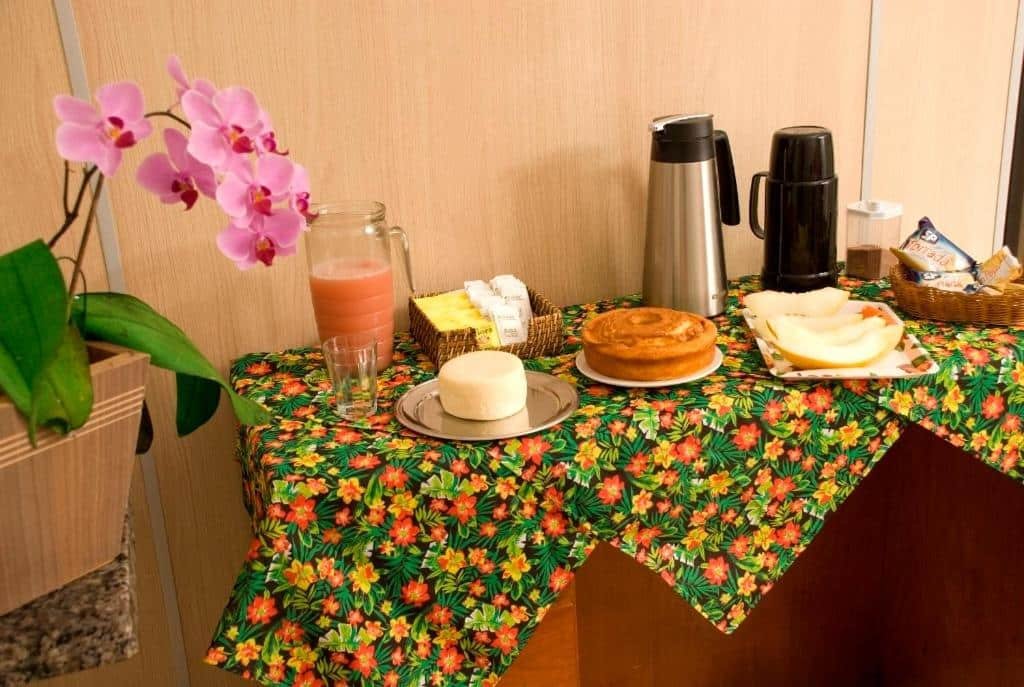 Café da manhã do Orquídea Hotel. No canto esquerdo há uma orquídea em uma balcão e no lado direito há uma pequena mesa com comidas e garrafas térmicas.