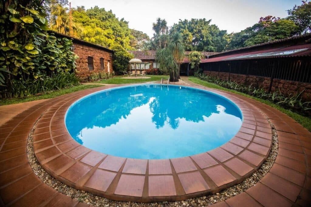 A imagem mostra uma piscina redonda de tamanho médio em um ambiente externo cercado por vegetação. A borda da piscina é feita de ladrilhos marrons, e ao redor há um caminho de pedras decorativas. Ao fundo, existem construções rústicas de pedra com janelas pequenas e telhados de barro, dando um toque histórico ao local. Algumas mesas e cadeiras estão dispostas sob um guarda-sol próximo à piscina. Árvores e plantas grandes cercam a área, criando um ambiente natural e tranquilo, perfeito para relaxar. Esse é o Paineiras Hotel Fazenda, um dos hotéis-fazenda em Minas Gerais.