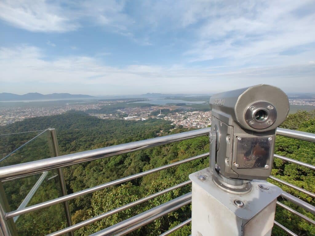 paisagem panorâmicas do Mirante de Joinville, mostrando todo o vale e a baía de Babitonga, com bastante vegetação natural, lagos, construções e um pequeno observador de metal no canto direito.
