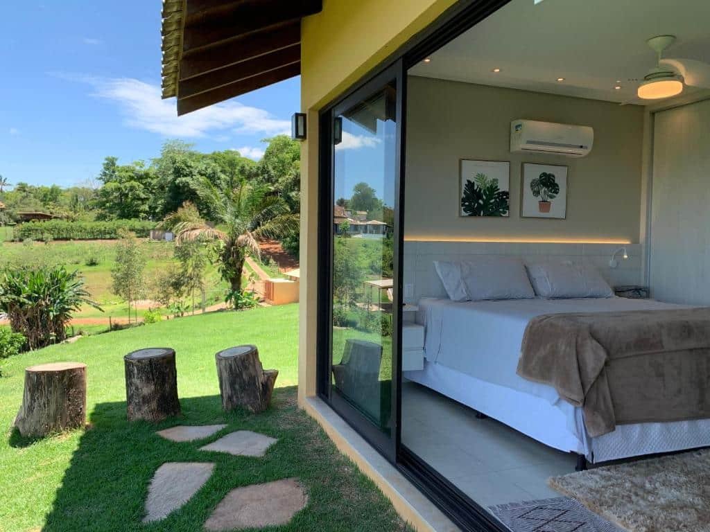 quarto com portas amplas de vidro abertas com vista para a cama de casal à direita e à esquerda, um campo gramado com bastante àrvores e vegetação natural