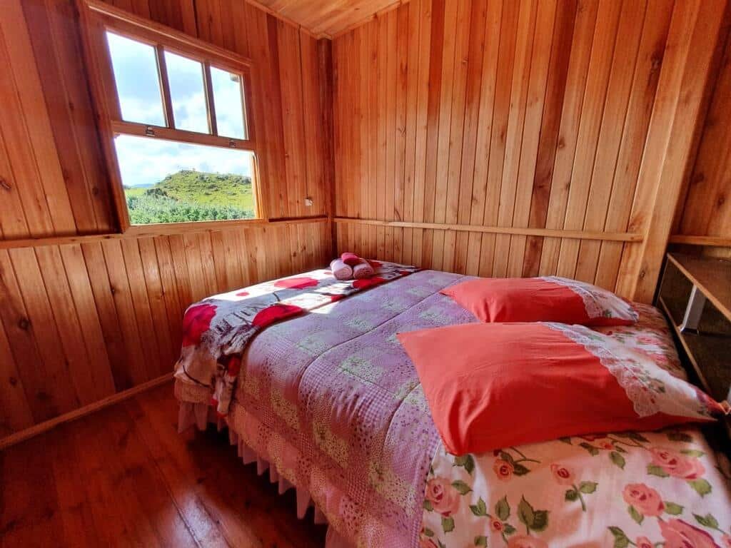Quarto do Paraíso Dos Cânions, com cama de casal em um ambiente simples todo em paredes de madeira. A roupa de cama é bastante colorida em retalhos. Há uma janela bem frente à cama em que é possível ver a natureza.