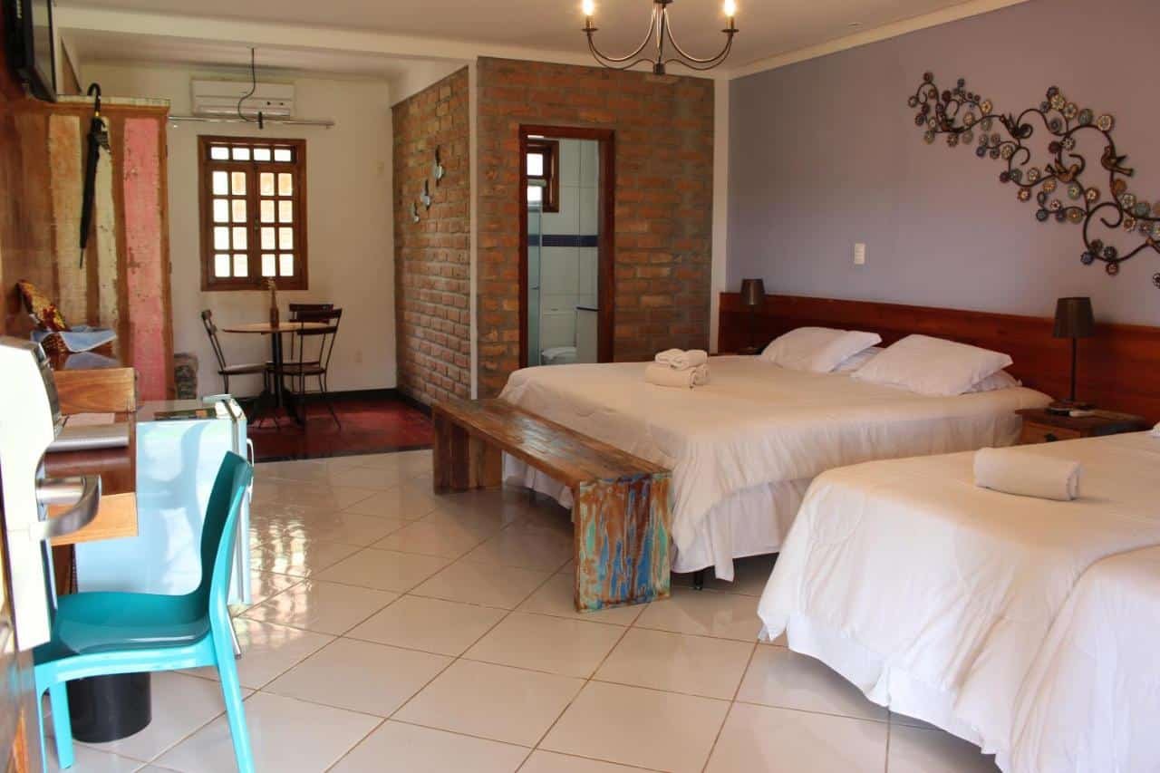 A imagem mostra um quarto espaçoso com duas camas de casal, ambas com lençóis brancos e travesseiros arrumados no Parque do Avestruz Eco Resort. Há uma decoração em ferro na parede acima da cama principal, e uma luminária pendente no teto. O ambiente tem paredes de tijolos aparentes e uma pequena mesa com cadeiras ao fundo, próxima a uma janela de madeira com persianas. O piso é de cerâmica branca brilhante, e há uma cadeira de plástico azul perto de uma pequena escrivaninha, ao lado de um frigobar. O quarto é simples, mas acolhedor, com toques rústicos e modernos.