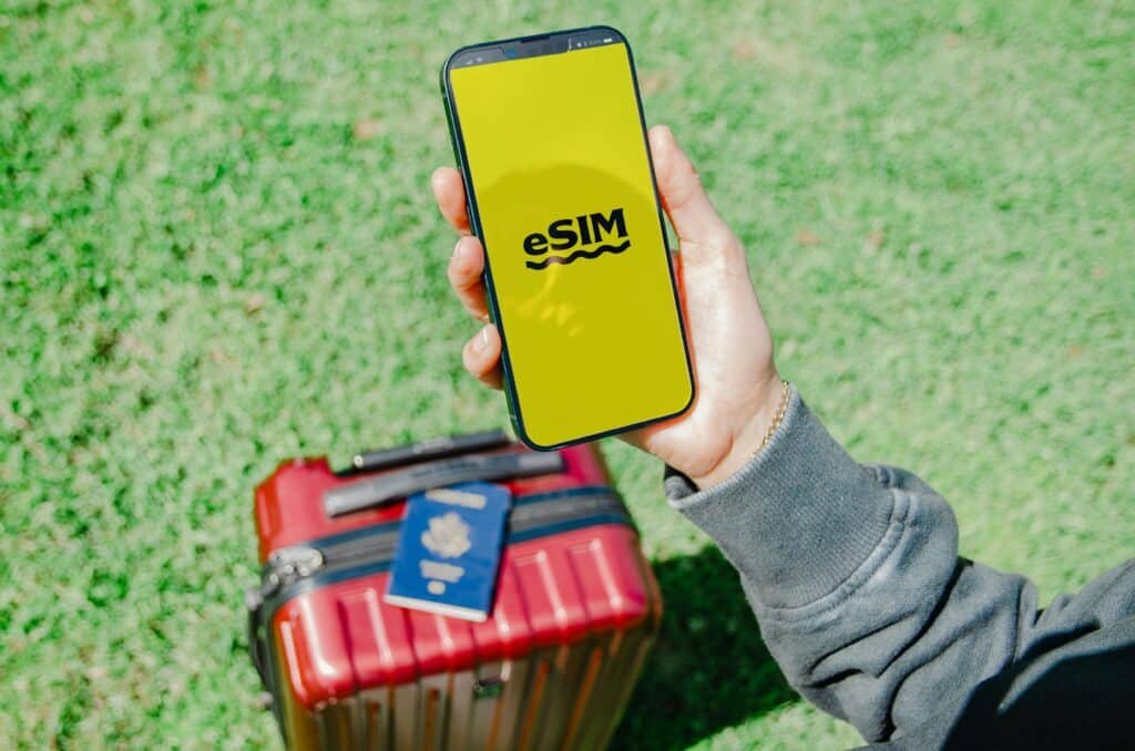 Uma pessoa segura um smartphone com a inscrição "eSIM" em uma tela amarela, acima de uma mala vermelha e um passaporte, na grama verde.