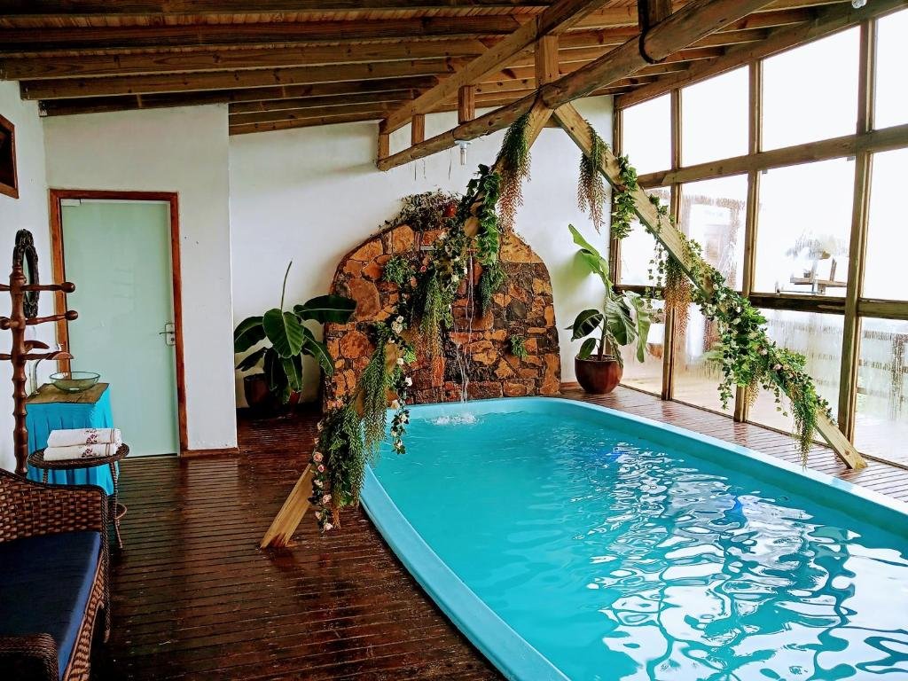 Piscina retangular com bordas arredondadas em um ambiente interno no Chalés da Serra Catarinense. Há uma espécie de arco acima da piscina com tufos de plantas decorativas. Do outro lado, a parede é totalmente de vidro com estrutura de madeira que dá vista para a natureza.