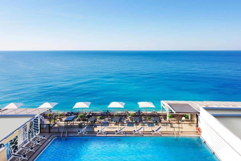 Cobertura do Le Meridien Nice. Há uma piscina grande retangular com uma grade de proteção, diversas mesas, espreguiçadeiras, guarda-sóis e uma vista privilegiada do mar.
