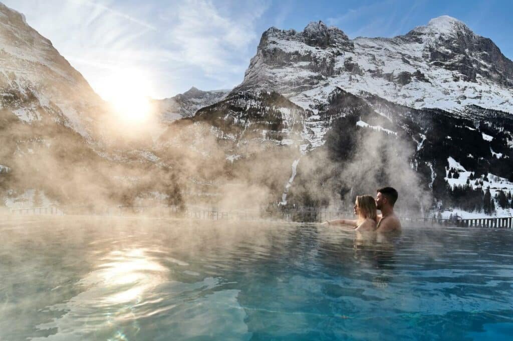 Imagem da piscina aquecida do Hotel Spinne Grindelwald durante o dia com um casal dentro da piscina do lado direito olhando as montanhas.