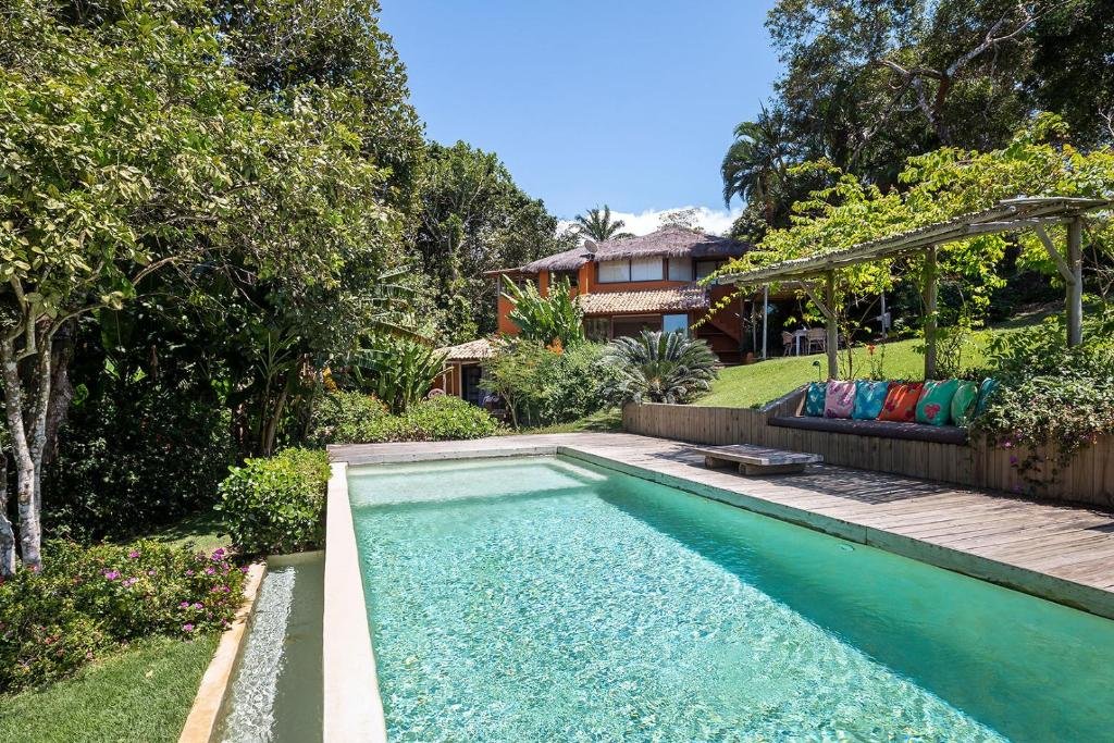 Piscina da Casas da Lia durante o dia, com árvores do lado esquerdo, com grama verde e ao fundo a hospedagem. Representa resorts em Trancoso.