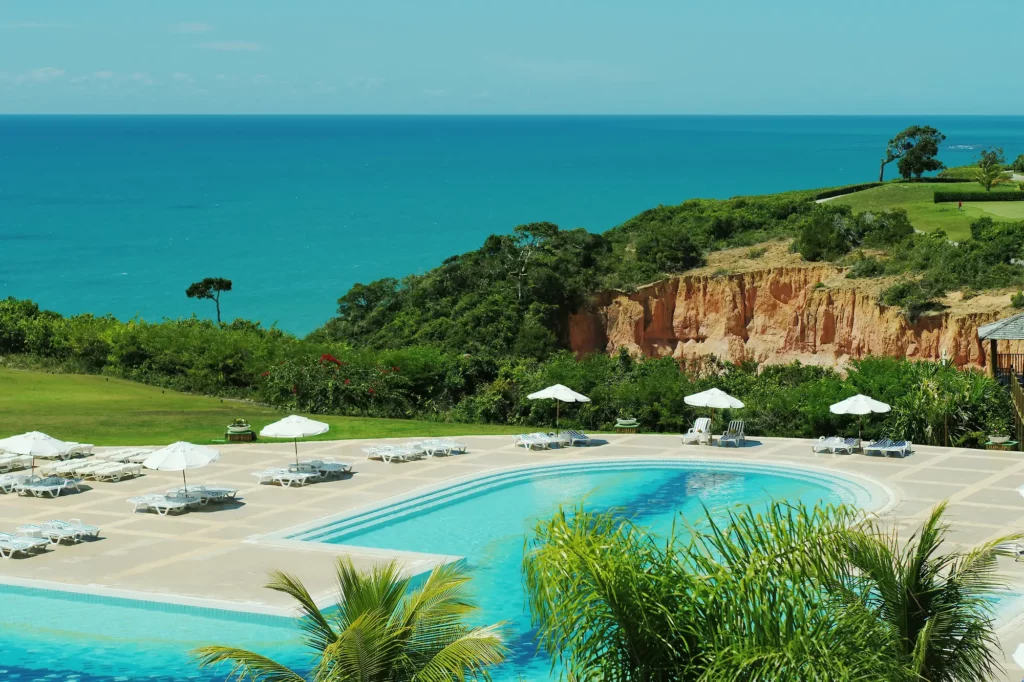 Vista da piscina do Club Med Trancoso durante o dia com cadeiras com guarda-sóis em volta e muita área verde. Representa resorts em Trancoso.