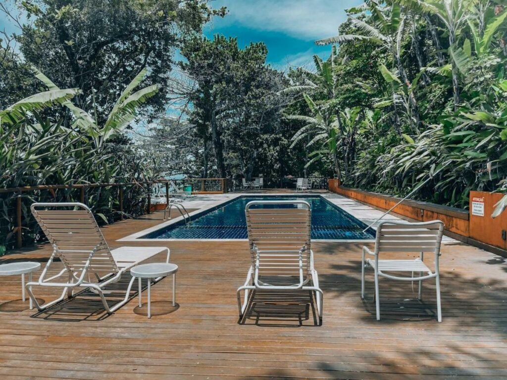 Imagem da piscina do Condomínio pé na areia – Santiago durante o dia com três cadeiras a frente e ao fundo a piscina.