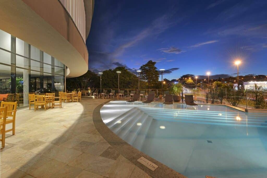 Grande piscina com várias mesas e cadeiras ao lado. Do lado de fora da propriedade é possível ver a rua. Está de noite.