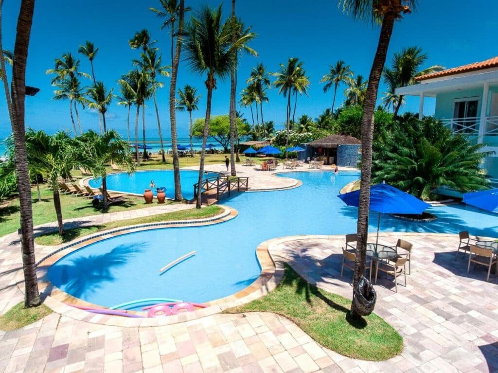 Área da piscina do Baía Branca Beach Resort, perto de Maragogi, com piscinas em formato redondo e várias palmeiras em volta. Há cadeiras com guarda-sóis espalhados e vista do mar ao fundo
